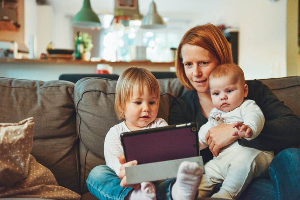 A person with short red hair is sitting on a grey couch with a baby in their lap and small child sitting next to them. They are looking at a purple iPad together and smiling.