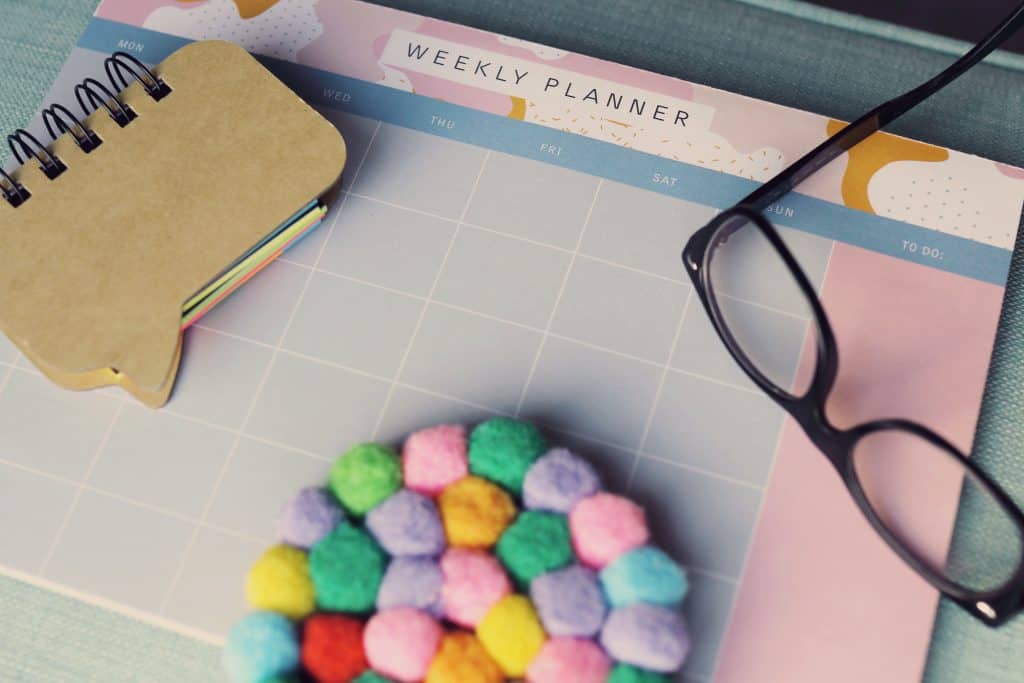 Close-up of a light purple and light pink weekly planner. A stack of brown notecards, a pair of black eye glasses, and a multicolor felt cup coaster are laying on top.