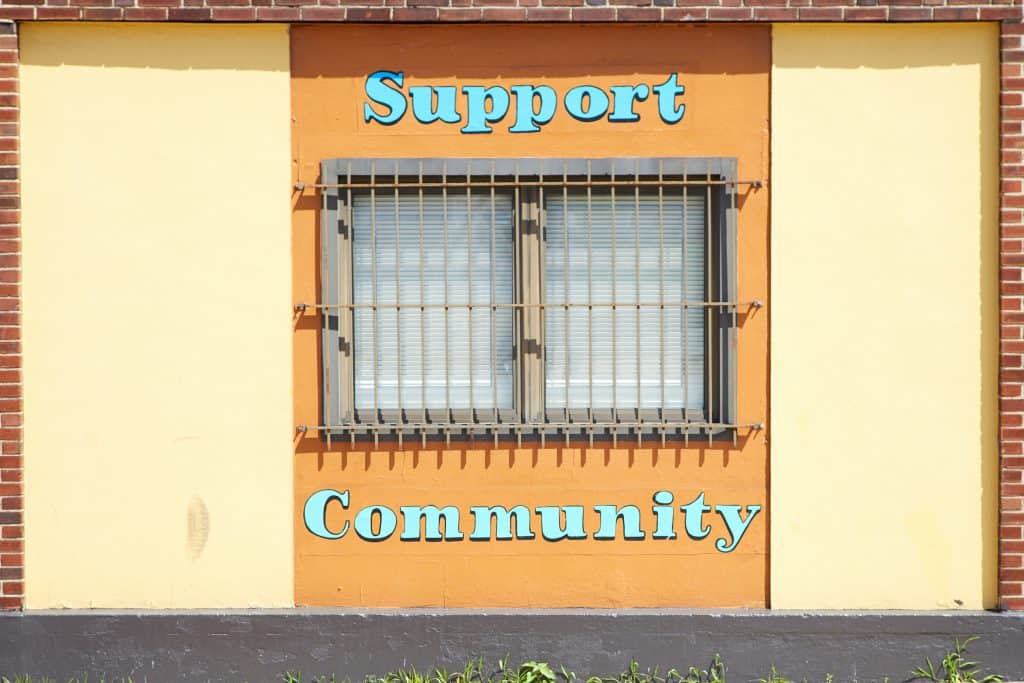 A yellow mural on the side of a building that says "Support Community". There is a window with bars on top that is between "Support" and "Community"