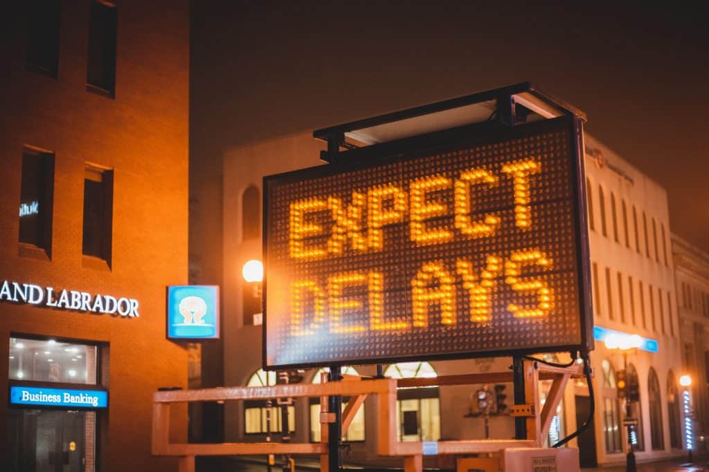A neon orange road sign that says "Expect Delays". It is night time.