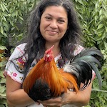 Julia is smiling in front of leaves and holds Fuego, a black and gold rooster