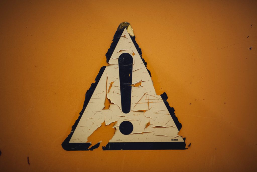 A photo of a light yellow triangle with a black exclamation point in the middle. The triangle is outlined in black. The background is dark orange.