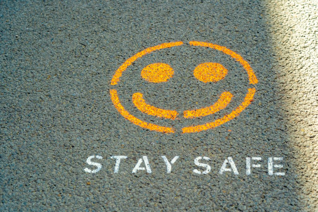 A photo of a stencil painting on the road. It's a painting of a dark yellow smiley face. Underneath, it says "Stay safe" in white capital painted letters.