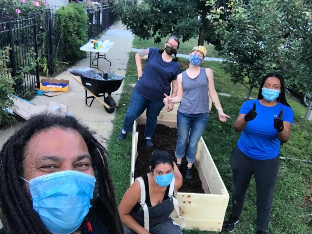 A photograph of five people wearing masks and giving thumbs up to the camera. They are standing around a newly-built garden bed that has soil in it. In the background, there are lots of trees and plants. On the left side of the photograph, there is a sidewalk that runs along a black rod iron fence. There is a white card table and a wheelbarrow in the middle of the sidewalk.
