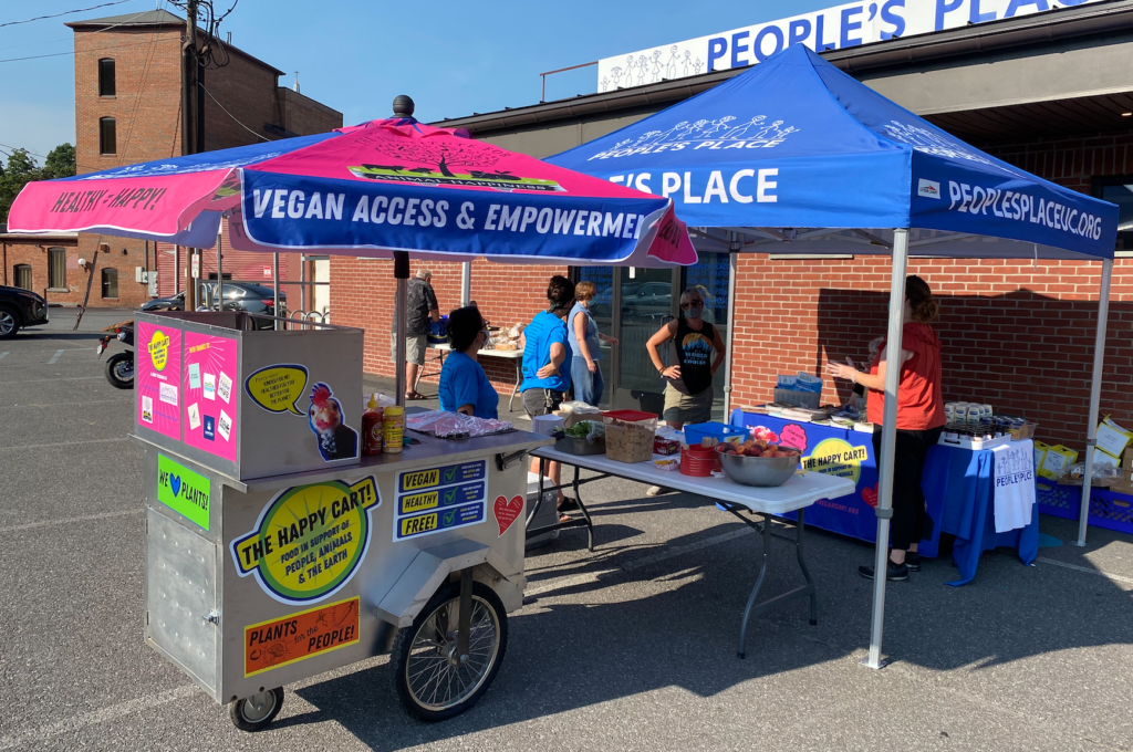A photograph of a silver food cart with lots of large colorful stickers on it. One reads "The Happy Cart!". A blue and pink umbrella is attached to the top of the cart. The umbrella has text on the side that reads "Vegan Access and Empowerment". To the right of the cart is a blue tent with a card table and four people standing underneath. There is food on the card table. The cart and the people are positioned on top of a parking lot and there is a brick building in the background with a sign on top that says "People's Place".