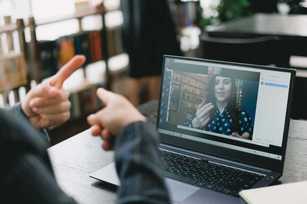 Photograph of an open laptop with one person onscreen and another person sitting in front of the laptop. They are on a video conferencing call utilizing sign language to communicate with one another.