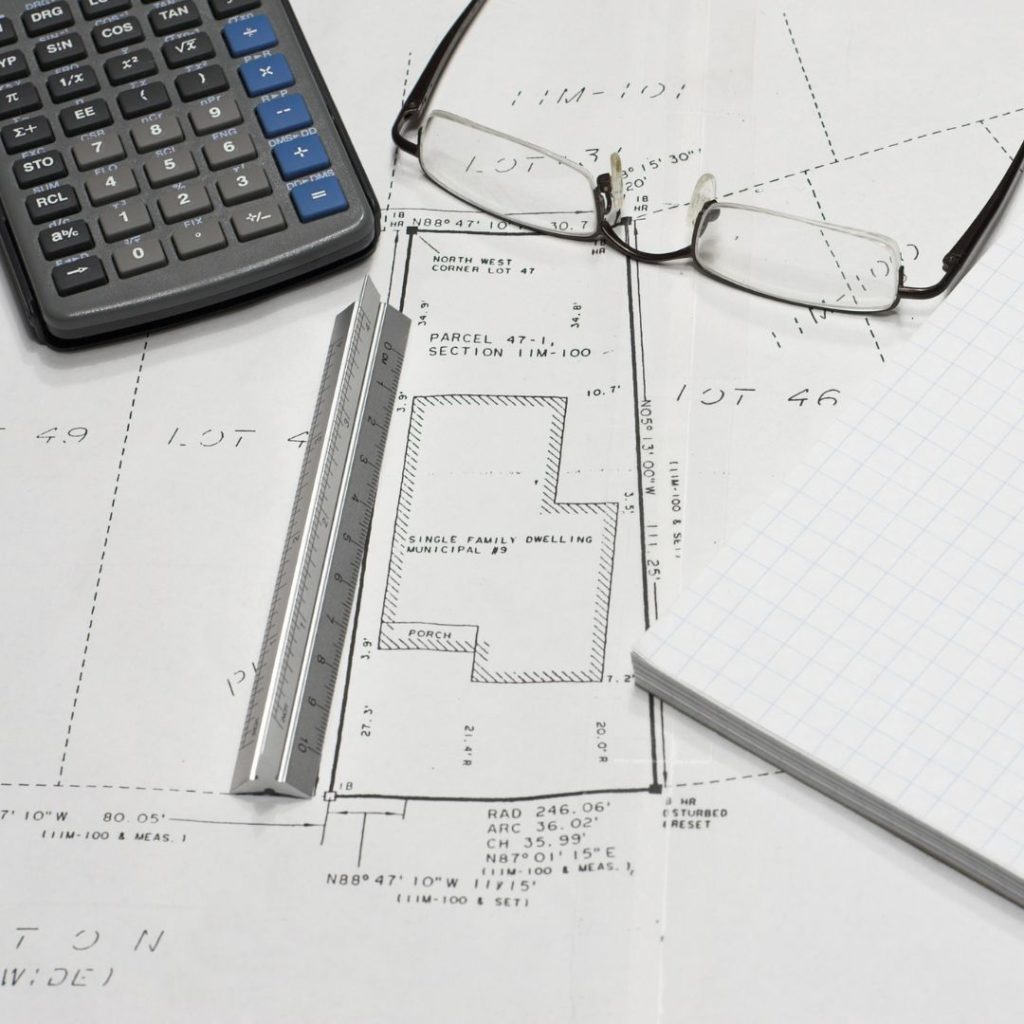 An image of a survey, showing a depiction of a square shaped piece of property along with lot line measurements, as well as where the building on the lot. Lying on the survey are a ruler, a calculator, eyeglasses, and graph paper.