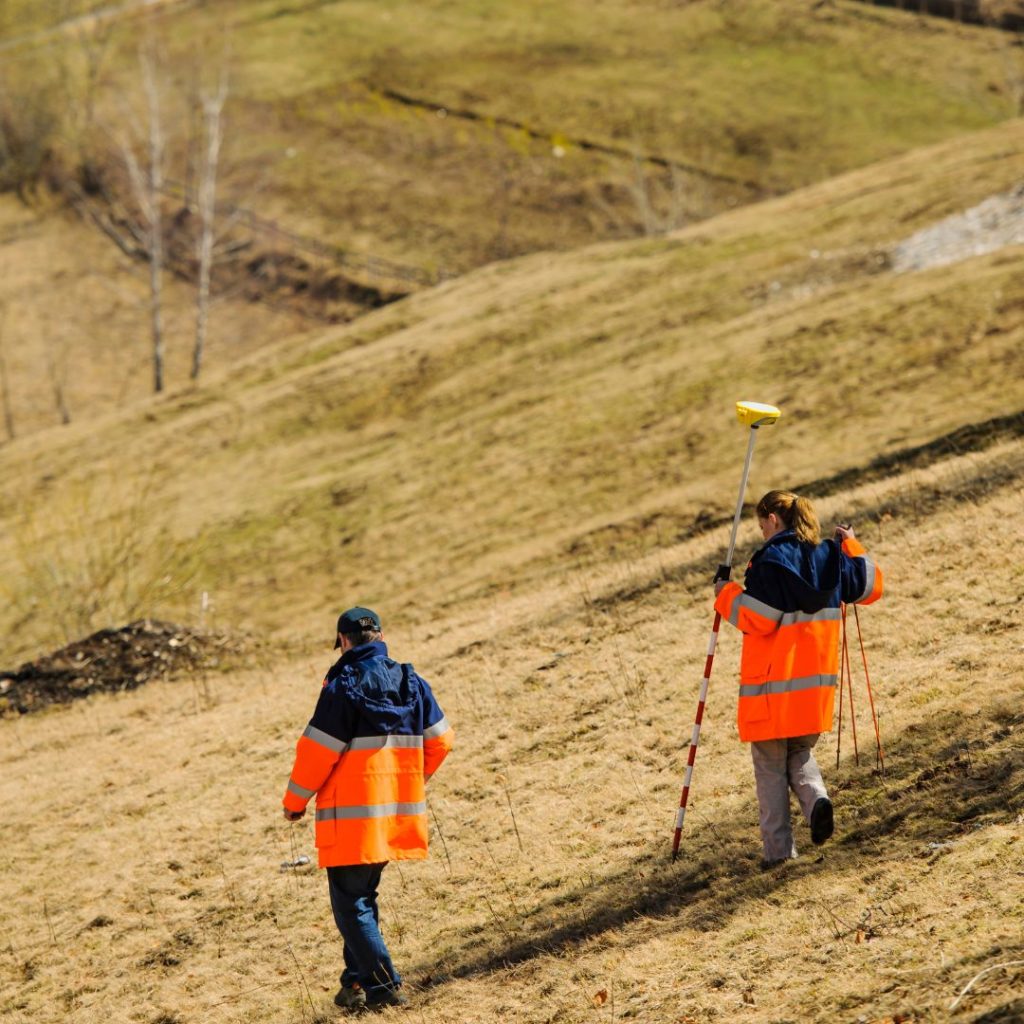 An image of two people in orange jackets walking across a hilly field carrying surveying equipment.