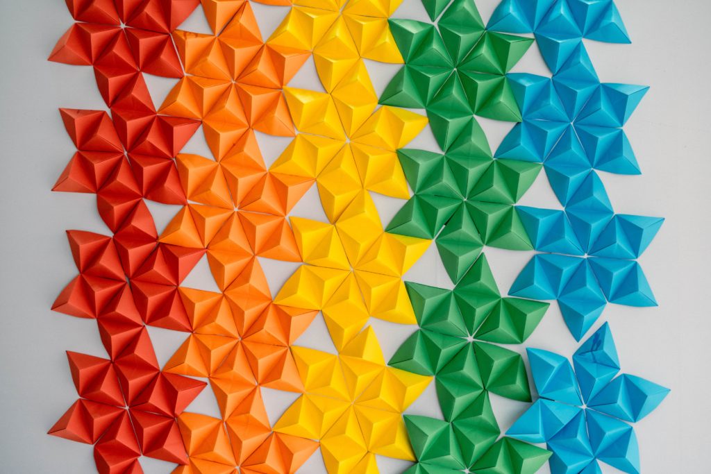 A close up photograph of an artwork made of multicolored origami stars. There are five columns of stars with four stars in each. The first column is red, the second column is orange, the third column is yellow, the fourth column is green, and the fifth column is blue.