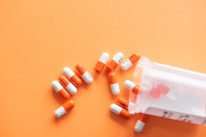 a clear prescription bottle lays on its side with orange and white capsules spilling out onto an orange background