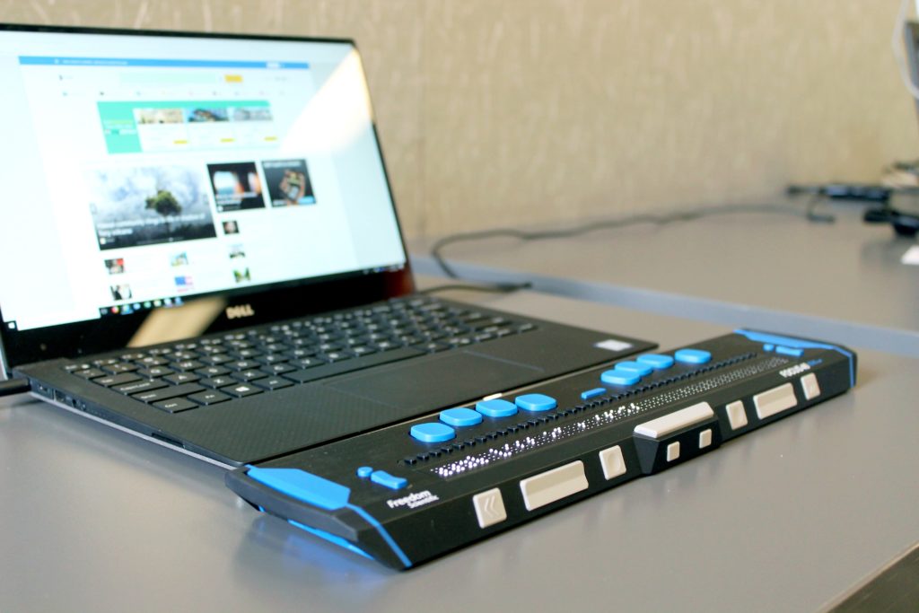 A close-up photograph of a black laptop with a refreshable braille display attached to it. The laptop is sitting on top of a grey table. The wall in the background is cream-colored.