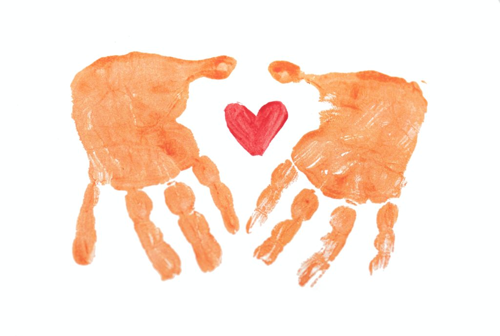Painted imprint of a pair of reddish-brown hands. There is a bright red heart painted in between the pair of hands.