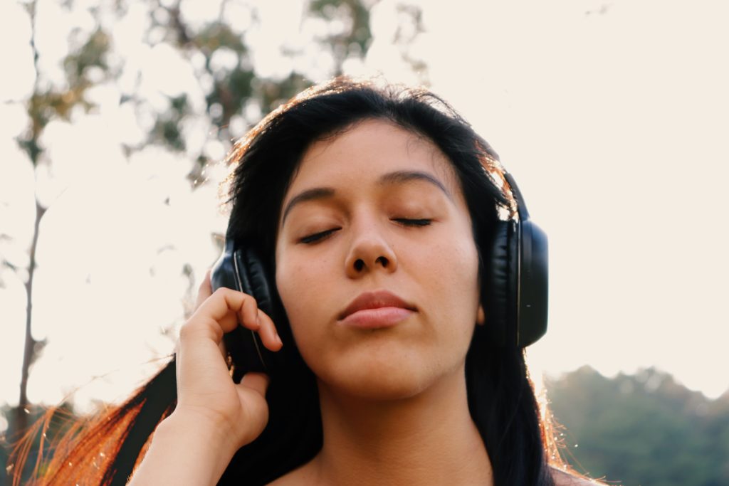 A close-up photograph of a person with long straight dark brown hair and light brown skin who is closing their eyes. They have black headphones on and they are touching the right side of the headphones with their right hand. 