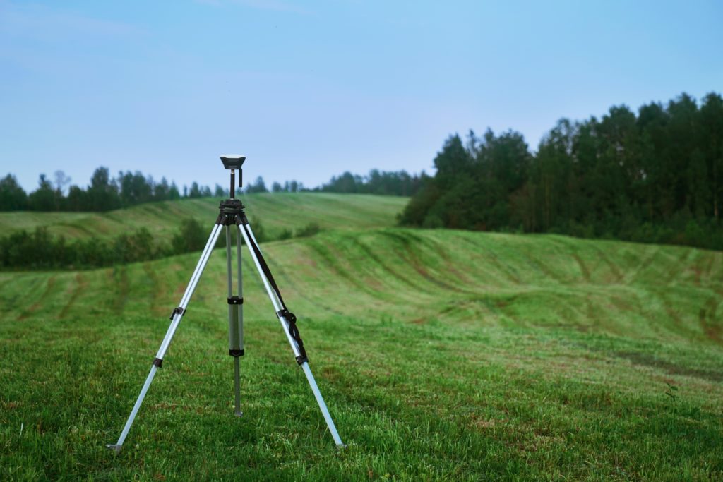 An image of surveying equipment standing in a green field with hills and trees.