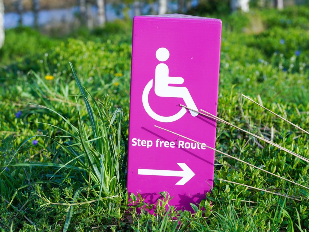A close-up photograph of a bright magenta sign sticking out of the grass. The sign has a white pictograph of the wheelchair symbol along with the words "step free route" written in white below. Below the words is a white arrow pointing to the right.
