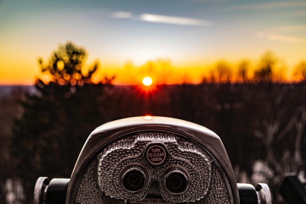 An image of binoculars facing towards a landscape with a sunrise. On the binoculars are the words "turn to clear vision."