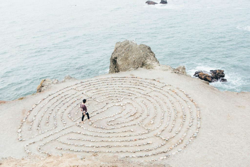 A person is walking through a labyrinth made of rocks on the beach. In the background, there is a large rock at the edge of the beach that extends out into a large body of water.