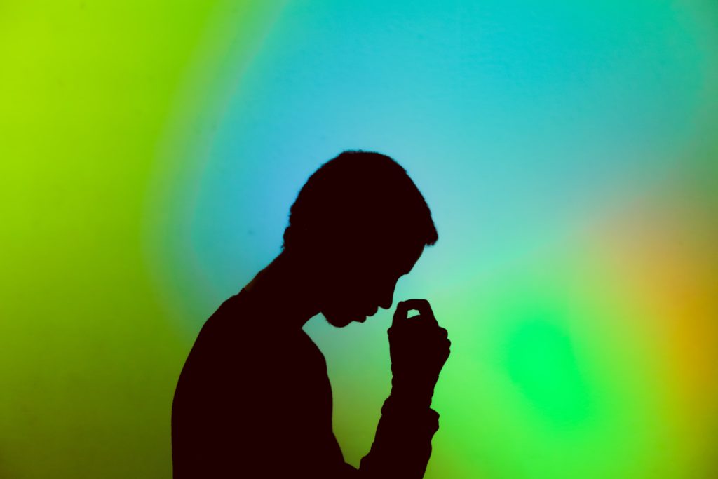 There is a shadowy person in the forefront of this image who is tilting there head downwards as if they are about to lean on their hand. The background consists of bright green, blue, and yellow light.
