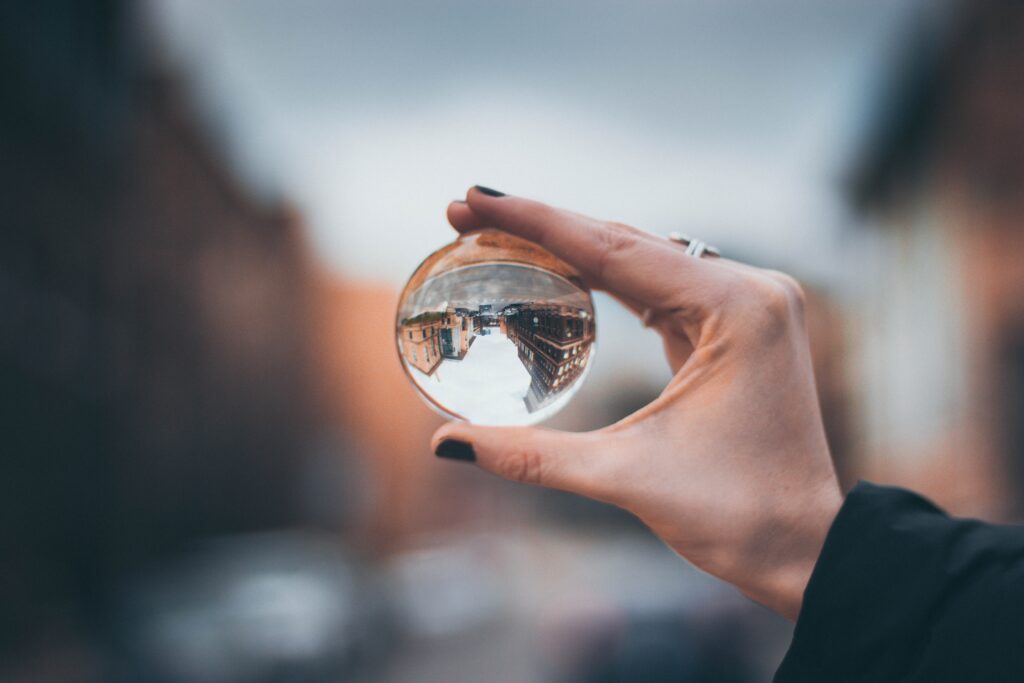 Photograph of a hand holding up a clear crystal ball. As you look through the ball, the scenery on the other side is distorted and upside down.