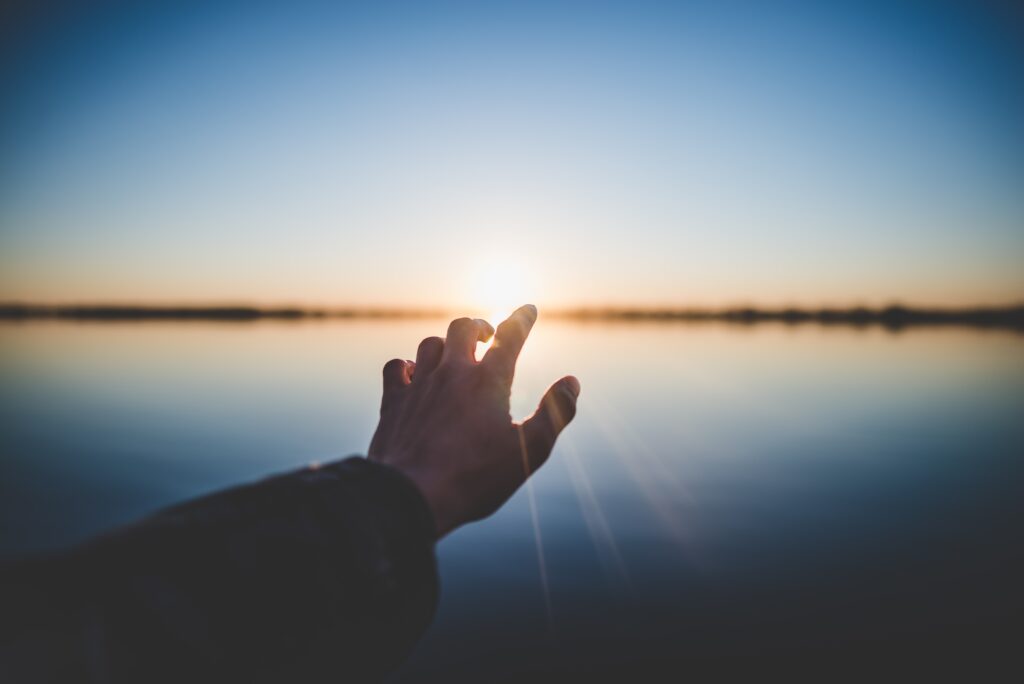 Photograph of a person's hand reaching out over a body of water toward the sunshine.