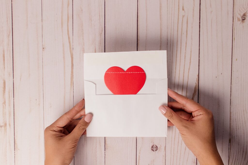 A photograph of a pair of hands holding an open envelope with a red paper heart inside. The background is a wood-paneled wall.