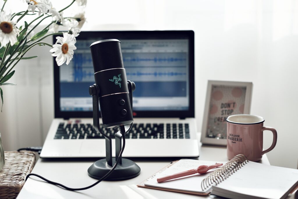 A photograph of a podcast microphone sitting on a desk in front of an open laptop. To the left of the microphone is a bouquet of white and yellow flowers. To the right of the microphone is a pink coffee cup, an open notebook with a pink pen on top, and a small white picture frame.