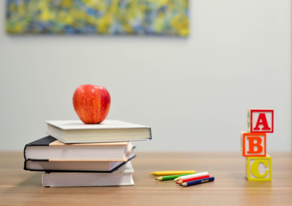 An image of a stack of books with an apple on top, some pencils, and some blocks printed with A, B and C on top of a desk. 