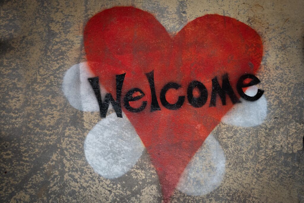 A photograph of a red heart that was spray painted onto concrete. There is a white flower that was spray painted behind the heart. There is also the word welcome on top of the red heart that was spray painted in black.