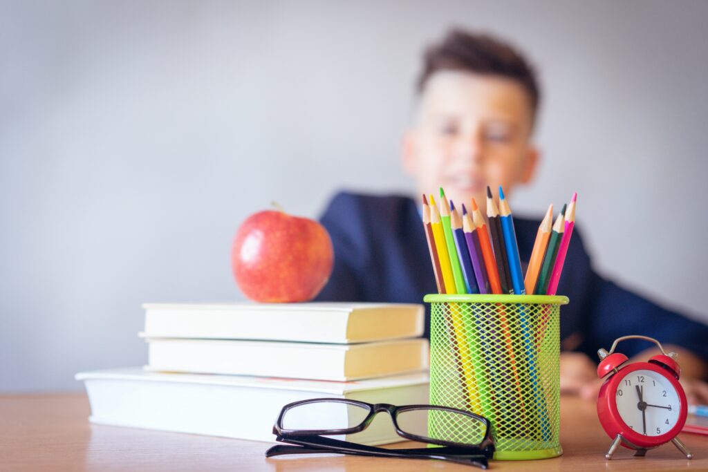 Close-up photograph of a stack of white books with a red apple on top. To the right of the books, there is a green pencil jar filled with colored pencils. There is a pair of black glasses sitting on the table in front of the pencil jar. There is also a small red clock sitting on the table to the right of the pencil jar. In the background, there is a blurred-out child who is smiling and wearing a dark blue shirt.