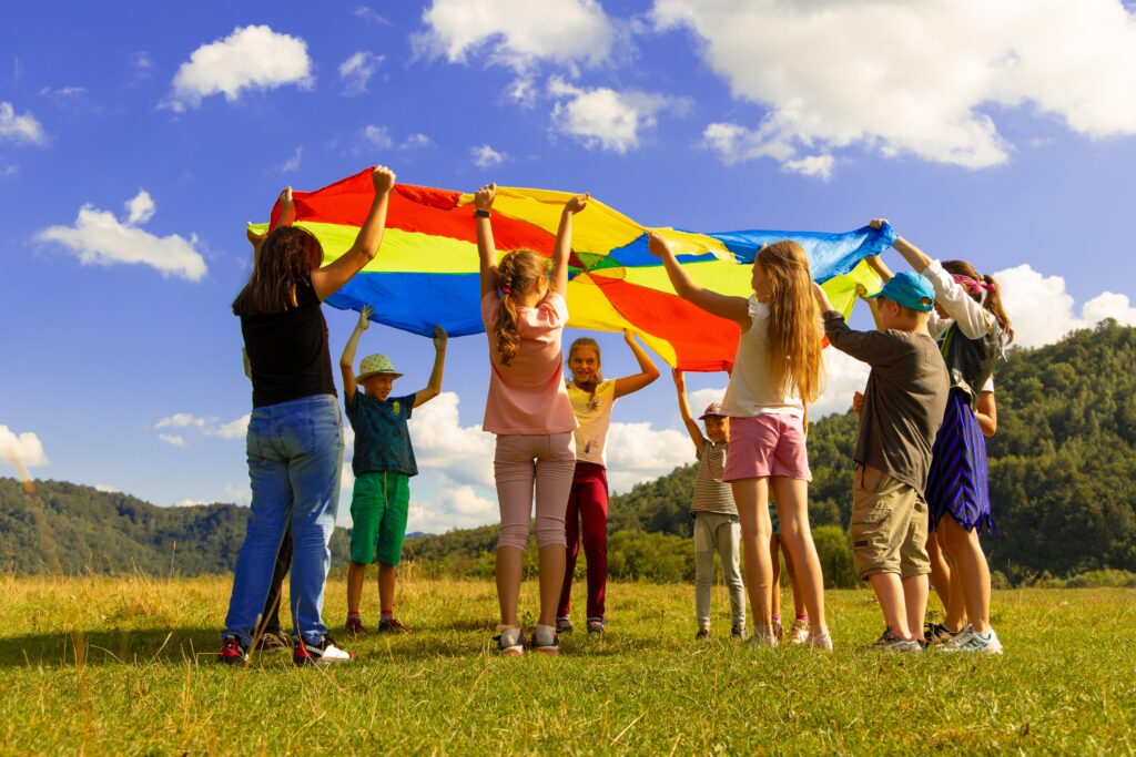 Photograph of a group of children playing with a rainbow-colored parachute in an open field. The sky is blue with some white fluffy clouds. There are green trees in the background.