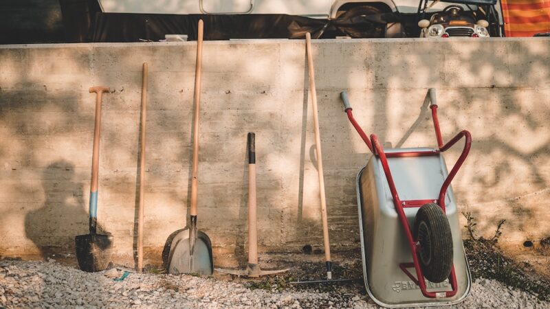 Shovels, rakes, pitchforks, a wheel barrow, and other tools lean against a wall.