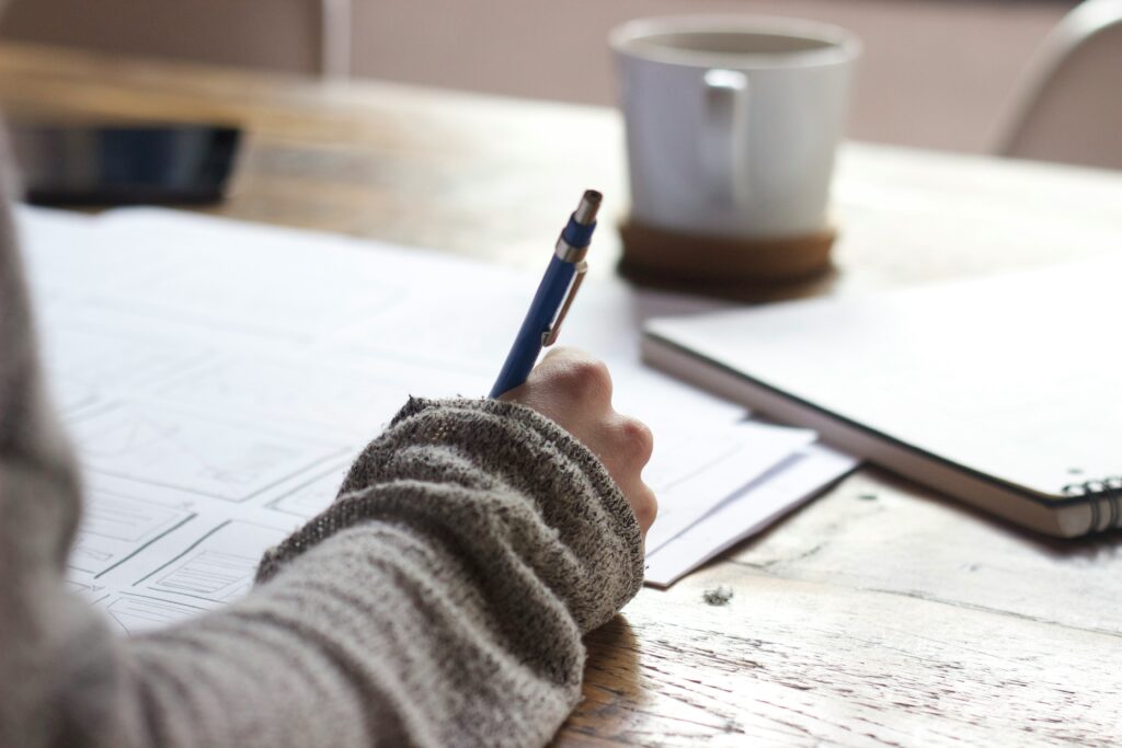 A photograph of a person's hand writing on a piece of paper. They are wearing a light brown sweater. There is a white coffee mug in the background.