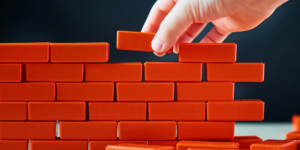 A hand places red toy bricks on top of one another, building a wall.
