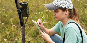 A caucasian personwith long brown hair pulled back in a ponytail crouches in a field making notes in a notebook. A trail camera is attached to a stick nearby.