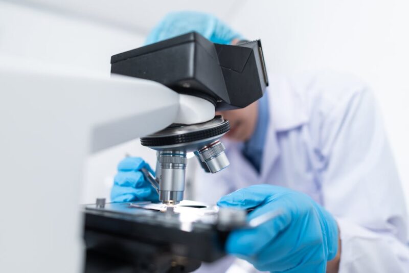 A person wearing a white lab coat, exam gloves, and a surgical cap works at a microscope.