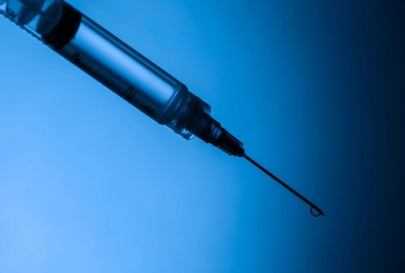 Liquid drips out of a needle attached to a syringe in front of a blue backdrop. 