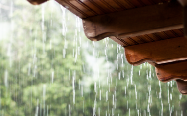 Rain pours past a wooden roof. Green trees can be seen through the rain in the distance.