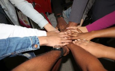 An image of eight hands reaching towards each other and clasping together at the center.