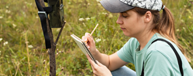 A caucasian personwith long brown hair pulled back in a ponytail crouches in a field making notes in a notebook. A trail camera is attached to a stick nearby.