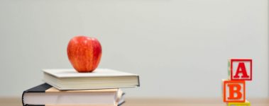 An image of a stack of books with an apple on top, some pencils, and some blocks printed with A, B and C on top of a desk.