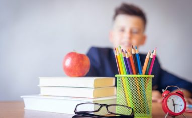 Close-up photograph of a stack of white books with a red apple on top. To the right of the books, there is a green pencil jar filled with colored pencils. There is a pair of black glasses sitting on the table in front of the pencil jar. There is also a small red clock sitting on the table to the right of the pencil jar. In the background, there is a blurred-out child who is smiling and wearing a dark blue shirt.